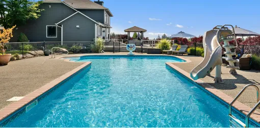 A large swimming pool surrounded by chairs and umbrellas in a backyard.